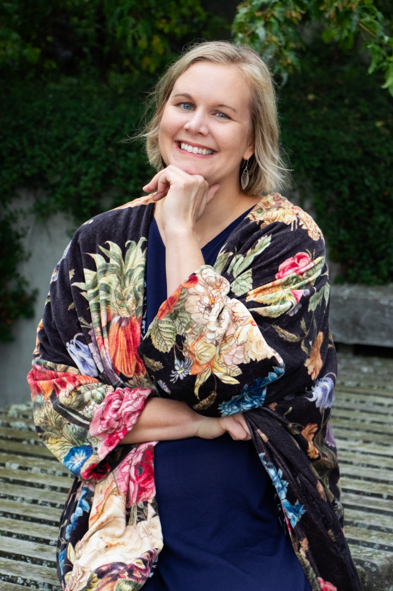 Psykoterapeutti Jonna Seppälä istuu ulkona, käsi leuan alla, kukkakuvioinen viitta ja taustalla vihreää kasvillisuutta.
